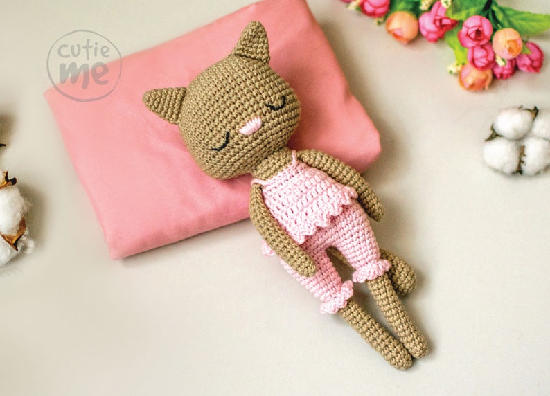 May include: A crocheted cat doll wearing pink pajamas, resting on a pink pillow. The doll has closed eyes, a pink nose, and is lying on its side. The image includes the text "cutie me" in a circle.