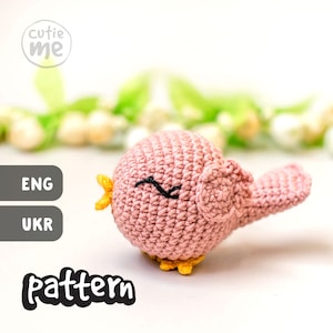 Peut inclure: Un oiseau en crochet rose avec des accents jaunes et des yeux noirs. L'oiseau est assis sur une surface blanche avec un fond floral vert et blanc. Le texte "cutie me" et "pattern" est visible sur l'image.