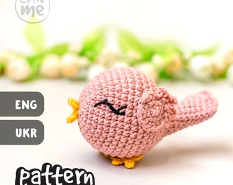 May include: A pink crocheted bird with yellow accents and black eyes. The bird is sitting on a white surface with a green and white floral background. The text "cutie me" and "pattern" are visible in the image.