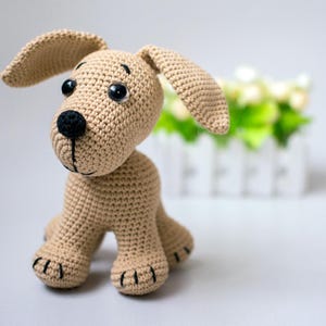 May include: A tan crocheted dog toy with large floppy ears, black eyes, and a black nose. The handmade stuffed animal has a stitched mouth and paws. The background includes a white picket fence and greenery.