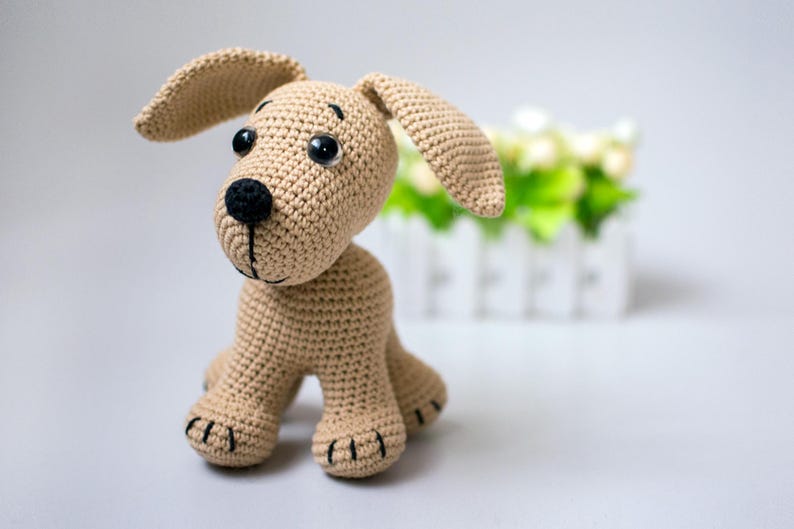 May include: A tan crocheted dog toy with large floppy ears, black eyes, and a black nose. The handmade stuffed animal has a stitched mouth and paws. The background includes a white picket fence and greenery.