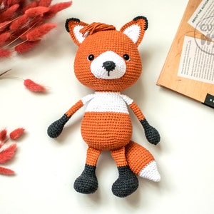 May include: A crocheted fox plush toy with orange and white fur, black eyes, and a black nose. The toy has black feet and a long, bushy tail.