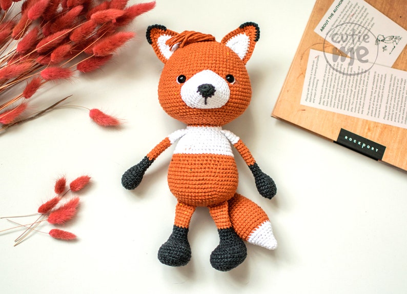 May include: A crocheted fox plush toy with orange and white fur, black eyes, and a black nose. The toy has black feet and a long, bushy tail.