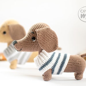 May include: Two crocheted dachshund dog plushies, one brown and one beige, both wearing striped sweaters. The brown dachshund is looking at the camera. The text "cutie me" is in a circle in the top right corner of the image.