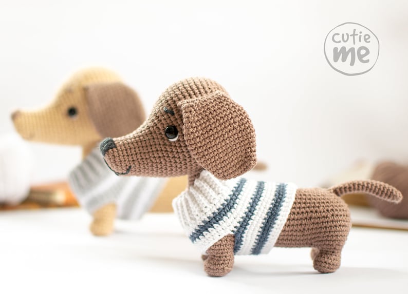 May include: Two crocheted dachshund dog plushies, one brown and one beige, both wearing striped sweaters. The brown dachshund is looking at the camera. The text "cutie me" is in a circle in the top right corner of the image.