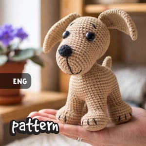 May include: A crocheted tan puppy with black eyes and nose, and floppy ears. The handmade dog is held in a person's hand. The word "pattern" is written in a white font at the bottom of the image, and the letters "ENG" are in a black box.
