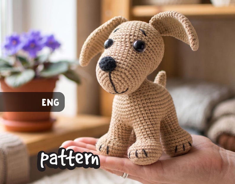 May include: A crocheted tan puppy with black eyes and nose, and floppy ears. The handmade dog is held in a person's hand. The word "pattern" is written in a white font at the bottom of the image, and the letters "ENG" are in a black box.