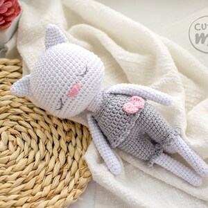 May include: A handmade crochet cat doll in a sleeping pose. The cat is primarily white with gray shorts and a pink bow. The cat's eyes are closed, and it has pink accents on its cheeks and ears. The doll is lying on a white cloth.