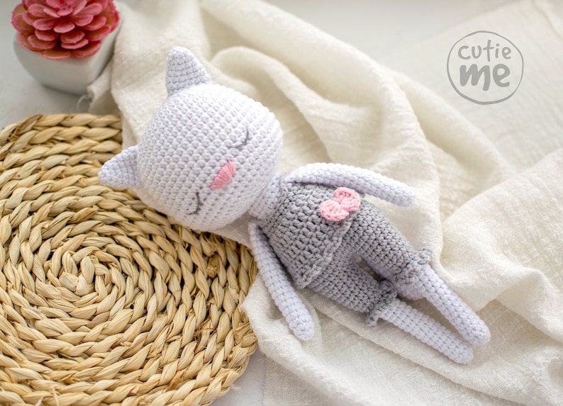 May include: A handmade crochet cat doll in a sleeping pose. The cat is primarily white with gray shorts and a pink bow. The cat's eyes are closed, and it has pink accents on its cheeks and ears. The doll is lying on a white cloth.