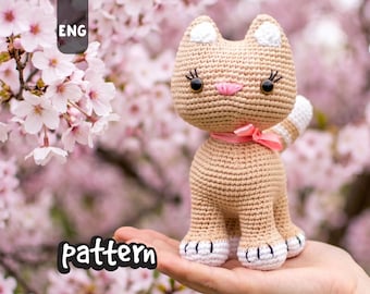 May include: A crocheted cat figurine in tan with white accents, a pink bow, and black eyes. The cat has a striped tail and is held in a hand. The word "pattern" is visible in the image.