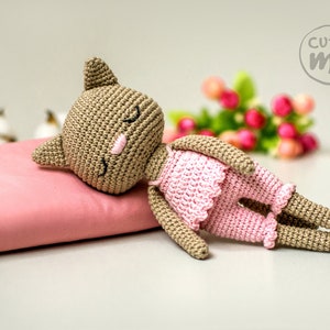 May include: A handmade crochet cat doll in a sleeping pose, wearing a pink dress and shorts. The doll is lying on a pink pillow. The doll has closed eyes and a pink nose. The image includes the text "cutie me".