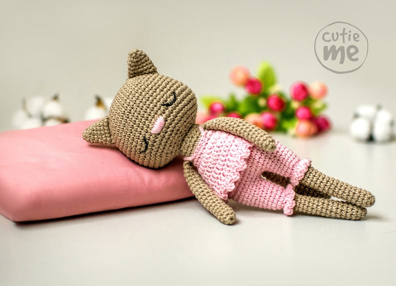 May include: A handmade crochet cat doll in a sleeping pose, wearing a pink dress and shorts. The doll is lying on a pink pillow. The doll has closed eyes and a pink nose. The image includes the text "cutie me".