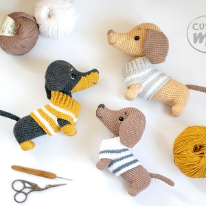 May include: Three crocheted dachshund dog plushies wearing striped sweaters. The plushies are brown, black and tan. There are also balls of yarn in the image.