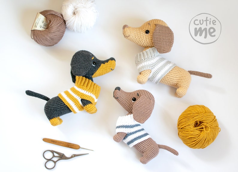 May include: Three crocheted dachshund dog plushies wearing striped sweaters. The plushies are brown, black and tan. There are also balls of yarn in the image.