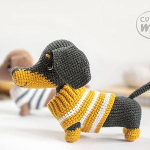May include: A crocheted dachshund wearing a yellow and white striped jumper. The dachshund is grey and black with a yellow nose and brown eyes. The dachshund is sitting on a white surface. The words "cutie me" are written in a circle in the top right corner of the image.