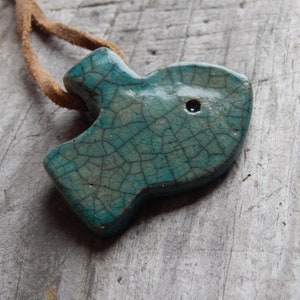 May include: A teal blue ceramic fish pendant with a crackled glaze and a small black hole. The pendant is attached to a brown leather cord.