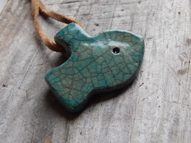 May include: A teal blue ceramic fish pendant with a crackled glaze and a small black hole. The pendant is attached to a brown leather cord.