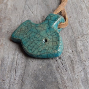 May include: A teal blue ceramic fish pendant with a crackled glaze finish. The pendant is attached to a brown leather cord.