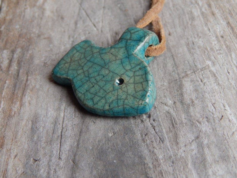 May include: A teal blue ceramic fish pendant with a crackled glaze finish. The pendant is attached to a brown leather cord.