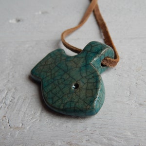 May include: A teal blue ceramic fish pendant with a crackled glaze finish. The pendant is attached to a brown leather cord.