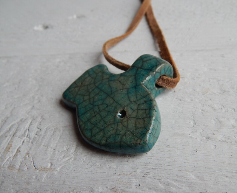 May include: A teal blue ceramic fish pendant with a crackled glaze finish. The pendant is attached to a brown leather cord.