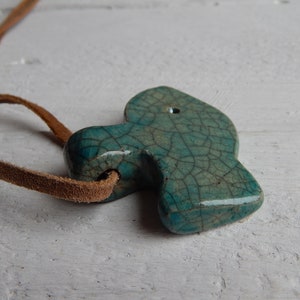 May include: A teal blue ceramic pendant with a crackled glaze finish, hanging from a brown leather cord. The pendant is shaped like a fish and has a hole at the top for hanging.