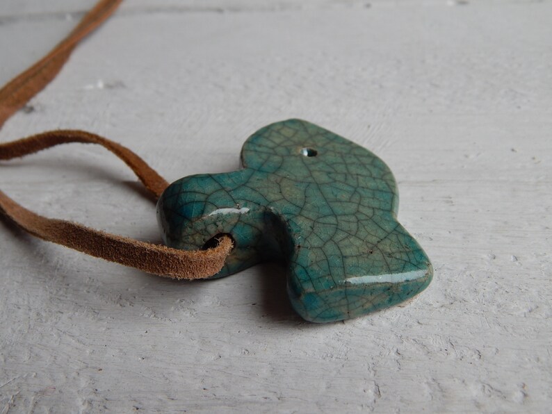 May include: A teal blue ceramic pendant with a crackled glaze finish, hanging from a brown leather cord. The pendant is shaped like a fish and has a hole at the top for hanging.