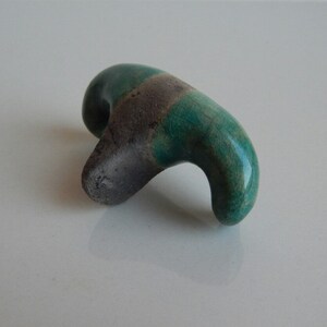 K&ouml;nnte beinhalten: Eine kleine, abstrakte Keramikskulptur mit einer gr&uuml;nen und grauen Glasur. Die Skulptur ist in Form eines Vogels oder Tieres mit einem langen, geschwungenen Hals geformt.