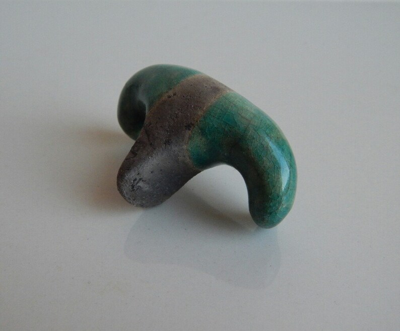 K&ouml;nnte beinhalten: Eine kleine, abstrakte Keramikskulptur mit einer gr&uuml;nen und grauen Glasur. Die Skulptur ist in Form eines Vogels oder Tieres mit einem langen, geschwungenen Hals geformt.