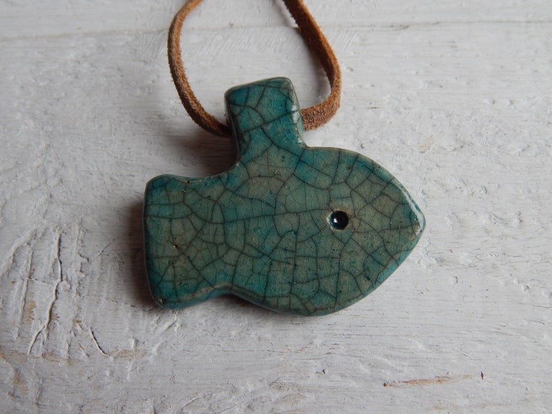 May include: A teal blue ceramic fish pendant with a crackled glaze and a black eye. The pendant is hanging from a brown leather cord.
