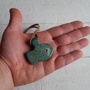 May include: A teal blue ceramic fish pendant with a crackled glaze finish. The pendant is hanging from a brown leather cord.