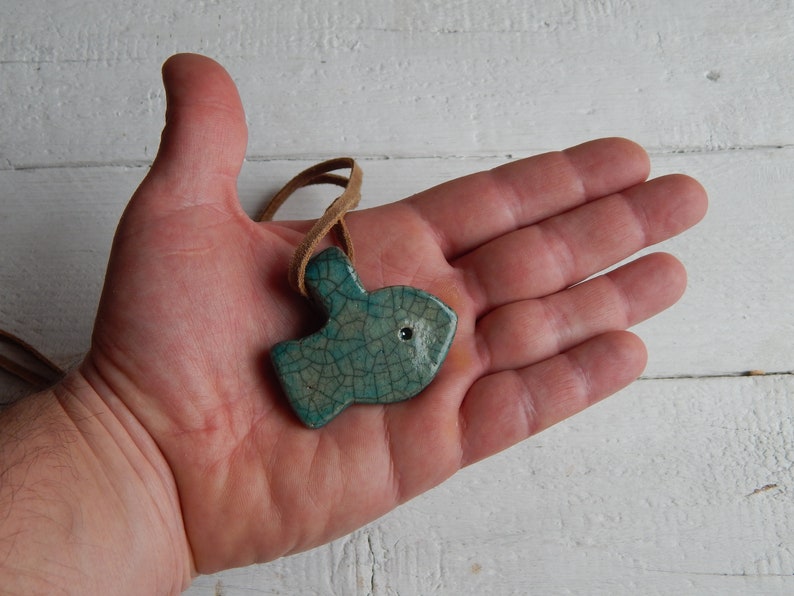 May include: A teal blue ceramic fish pendant with a crackled glaze finish. The pendant is hanging from a brown leather cord.