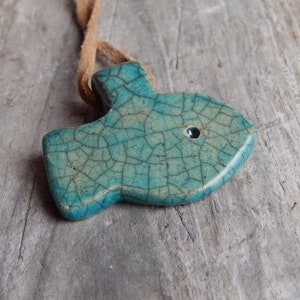 May include: A teal blue ceramic fish pendant with a crackled glaze and a black eye. The pendant is attached to a brown leather cord.