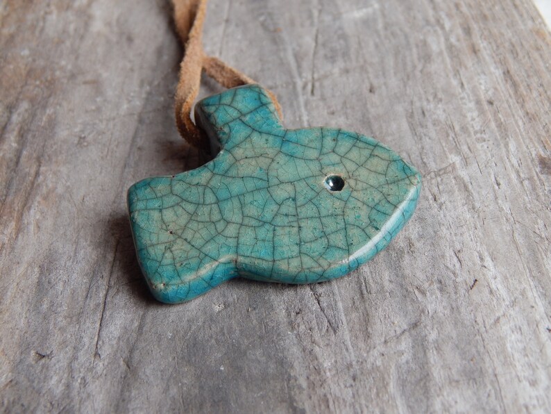 May include: A teal blue ceramic fish pendant with a crackled glaze and a black eye. The pendant is attached to a brown leather cord.