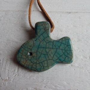 May include: A teal blue ceramic fish pendant with a crackled glaze finish. The pendant is attached to a brown leather cord.