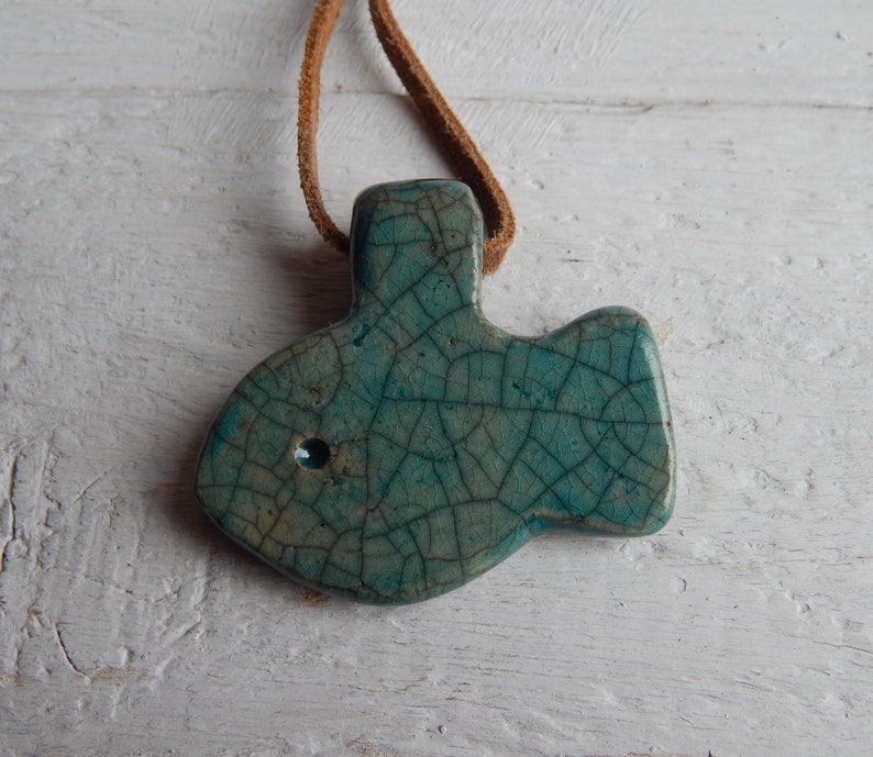 May include: A teal blue ceramic fish pendant with a crackled glaze finish. The pendant is attached to a brown leather cord.
