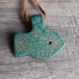 May include: A teal blue ceramic fish pendant with a crackled glaze and a small black eye. The pendant is attached to a brown leather cord.