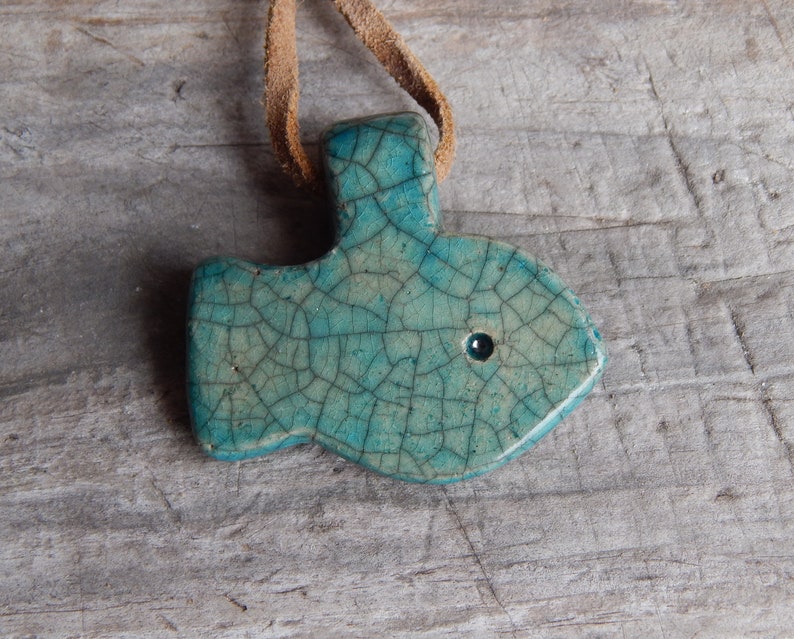 May include: A teal blue ceramic fish pendant with a crackled glaze and a small black eye. The pendant is attached to a brown leather cord.