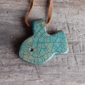 May include: A teal blue ceramic pendant shaped like a fish with a crackled glaze finish. The pendant is hanging from a brown leather cord.