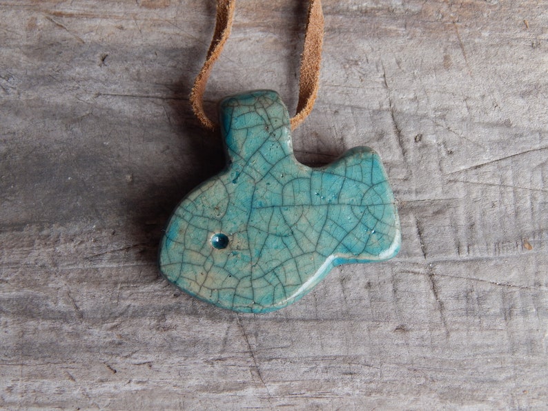 May include: A teal blue ceramic pendant shaped like a fish with a crackled glaze finish. The pendant is hanging from a brown leather cord.