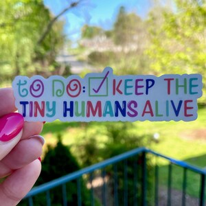 May include: A colorful sticker with the text "To Do: Keep the Tiny Humans Alive" with a checkmark in a box. The sticker is held in a hand with pink nail polish.