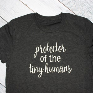 May include: A dark gray t-shirt with the text "protector of the tiny humans" printed in white.