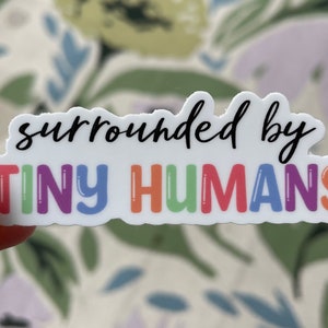 May include: A white sticker with the phrase "surrounded by TINY HUMANS" in black and rainbow lettering. The sticker is held by a person with a pink nail polish. The background has a floral pattern.