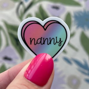 May include: A white heart-shaped sticker with a pink and blue ombre design. The word "nanny" is written in black inside the heart.