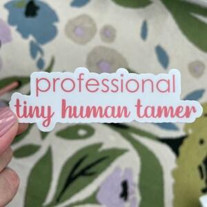 May include: A white sticker with the words "professional tiny human tamer" in pink script. The sticker is rectangular with rounded corners. The background is a floral pattern with blue, green, and white flowers.