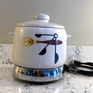 May include: A white ceramic slow cooker with a speckled finish and a lid. The front features a graphic illustration of a yellow fork and black measuring spoons. The pot sits on a circular chrome base with an attached black electrical power cord.