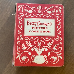 May include: A red hardcover cookbook with a white floral design and the title "Betty Crocker's Picture Cook Book" with the year 1950 printed below.