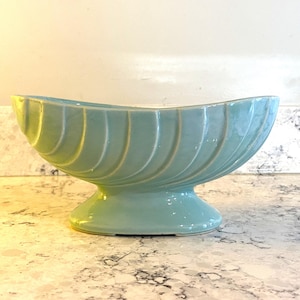May include: A light blue ceramic bowl with a curved, elongated oval shape and a pedestal base. The exterior features a series of vertical, raised ridges that wrap around the body, creating a textured, scalloped appearance with a smooth finish.
