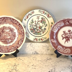 May include: Three ceramic plates displayed on black stands. The left plate features a maroon landscape scene with a scalloped edge. The center plate has a multi-colored floral tree design. The right plate displays a maroon floral motif on a white base.