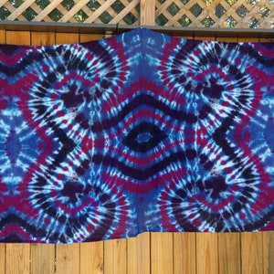 May include: A large tie-dye tapestry with a symmetrical pattern in shades of blue, purple, and red. The tapestry is hanging on a wooden fence.